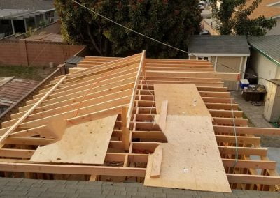 A wooden frame of a partially constructed roof on a house with surrounding trees and neighboring homes in the background, reminiscent of a modern California garage conversion project.